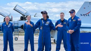 four astronauts up in blue flight suits stand behind microphones on a airplane runway. two white n' blue jets is behind them