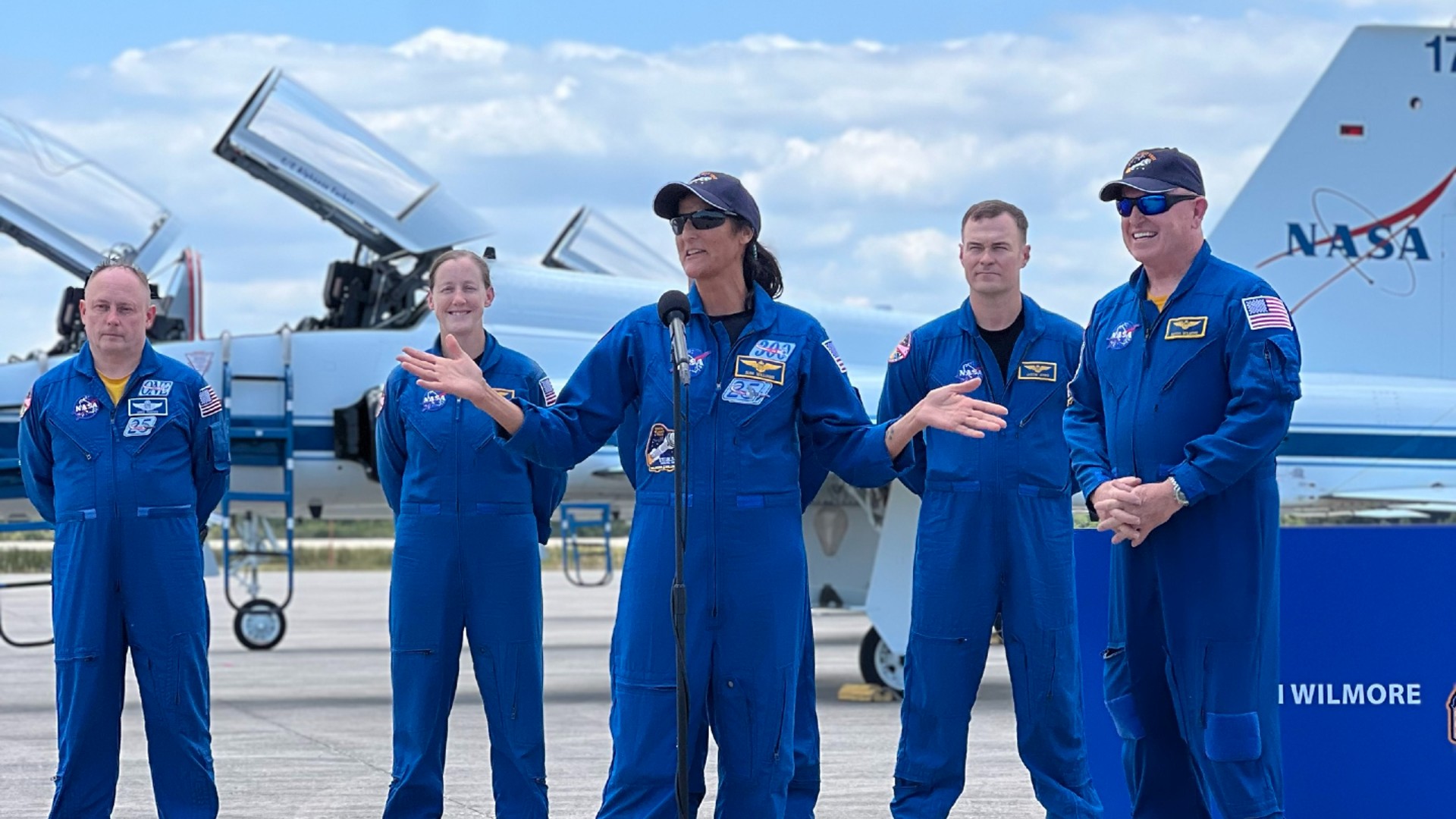 NASA Space Technology four astronauts in blue flight suits stand on the encourage of microphones on an airplane runway. two white and blue jets are on the encourage of them