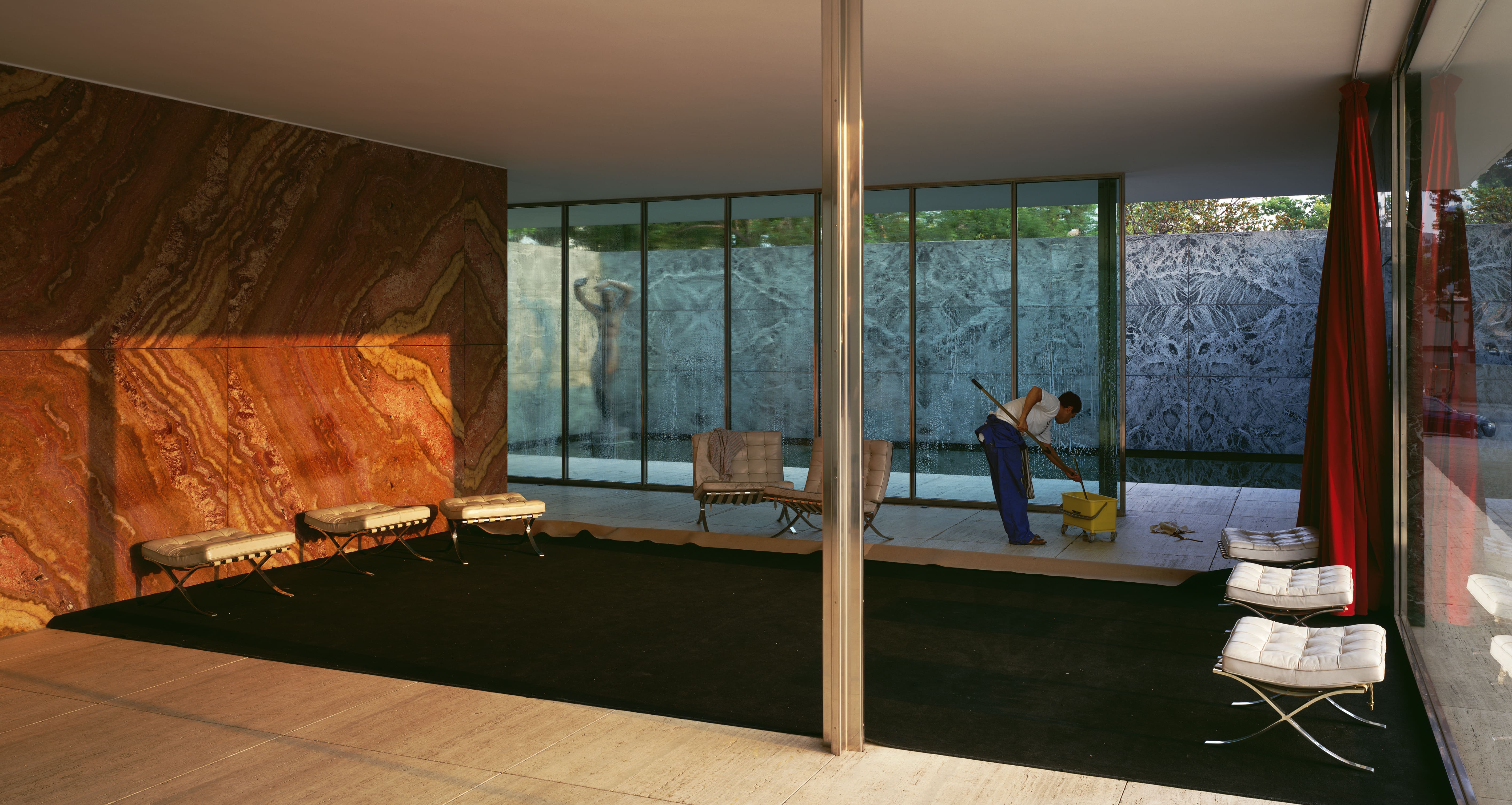 A worker mops the floor of a modernist glass and stone interior, possibly the Barcelona Pavilion, with sunlight streaming through the windows.