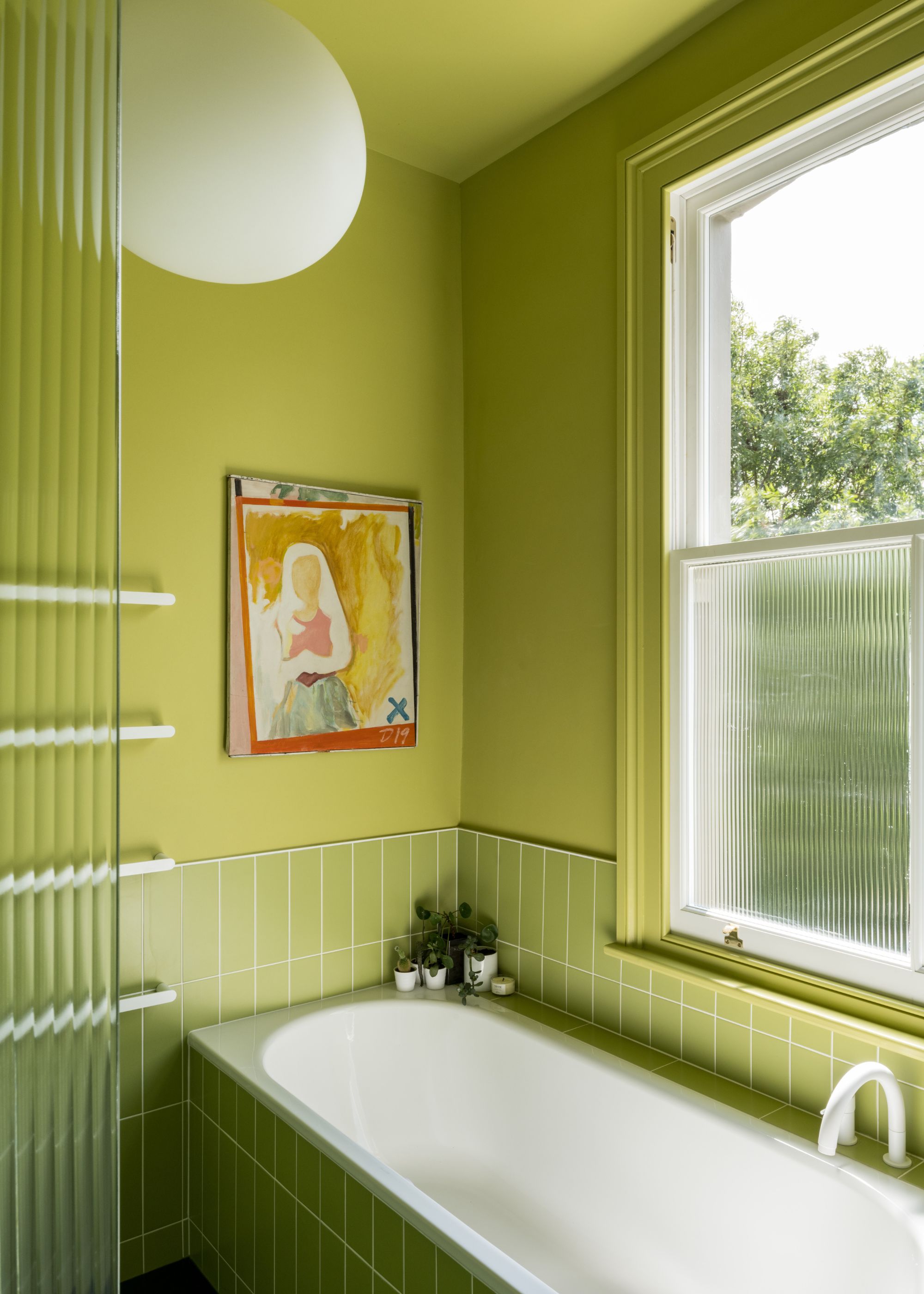 The corner of a bathroom with chartreuse walls and matching wall tiles, a sash window, and a white bathtub with green tiles on the outside.