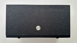 The front of the Audio Pro Addon C10 MkII wireless speaker, now with its magnetic griller attached. This is a gray fabric with a rough texture, and a silver circular logo near the bottom of the center.