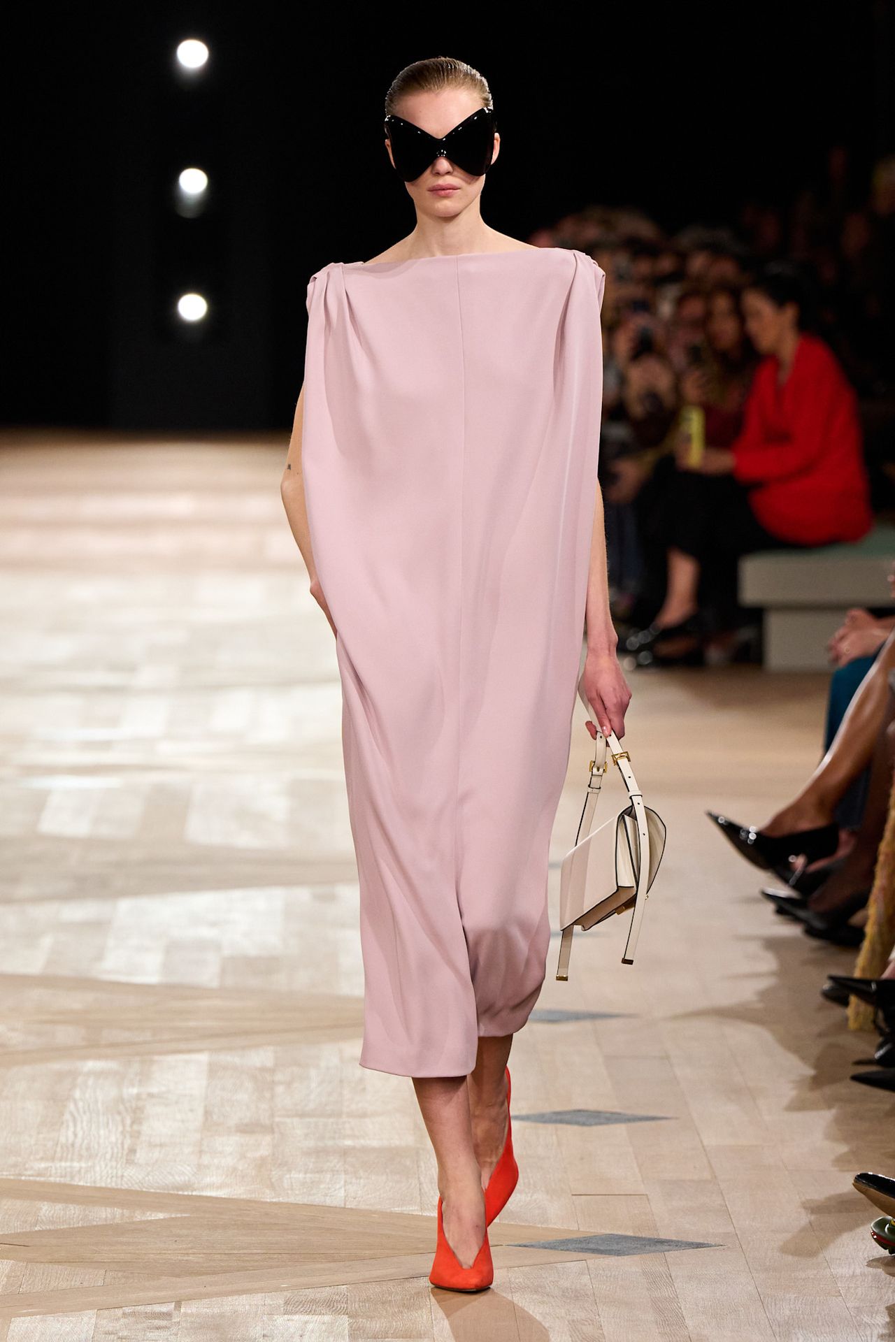 A Balenciaga model wearing a pale pink midi dress styled with pointed red pumps, a small white handbag, and oversize black bug-eye sunglasses.