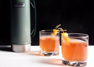two rocks glasses are filled with an orange cocktail. A Thermos sits nearby for storing the drinks before serving