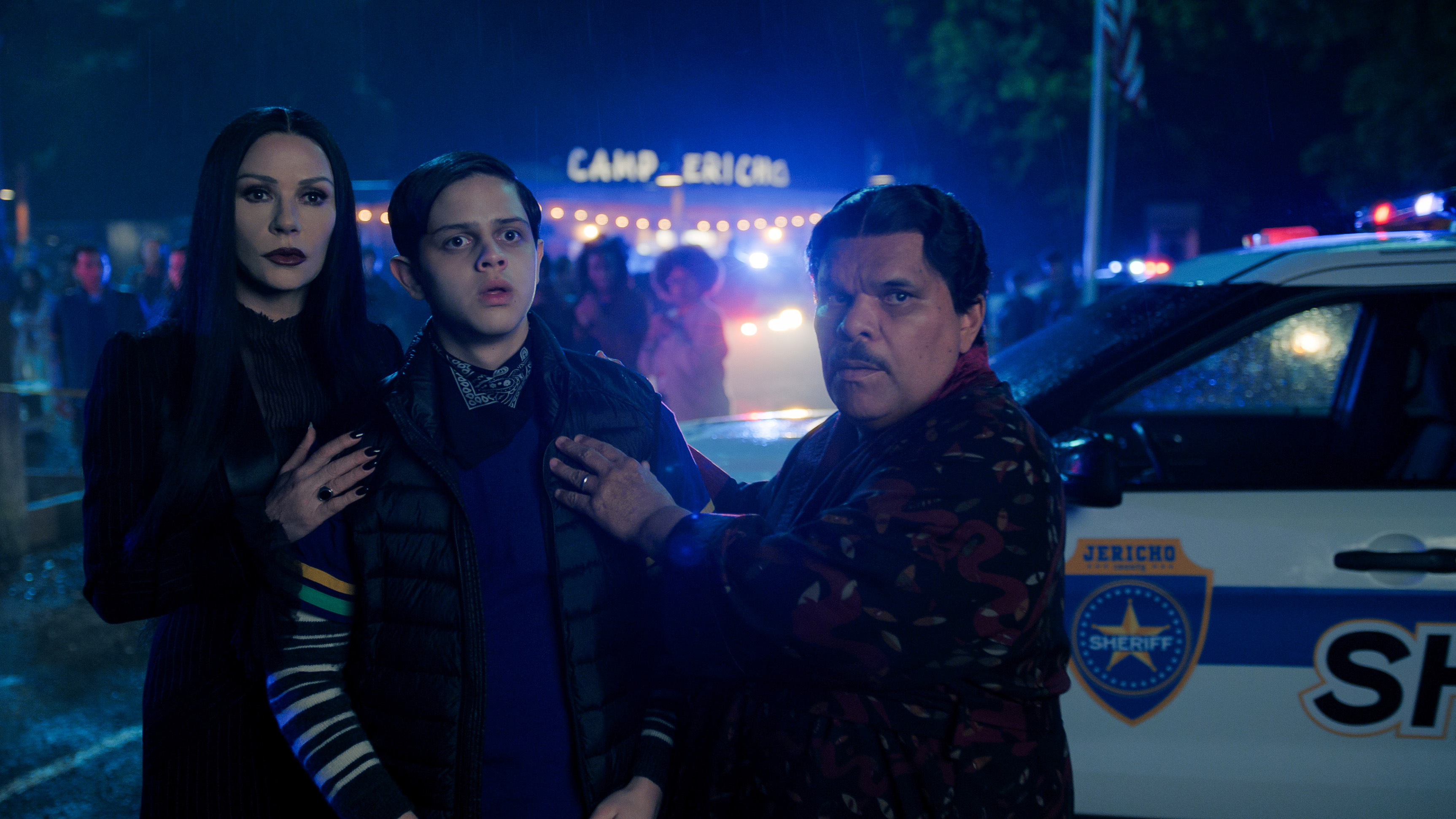 Catherine Zeta-Jones as Morticia Addams, Isaac Ordonez as Pugsley Addams, and Luis Guzma&amp;#769;n as Gomez Addams all stand near a police car in Wednesday season 2.