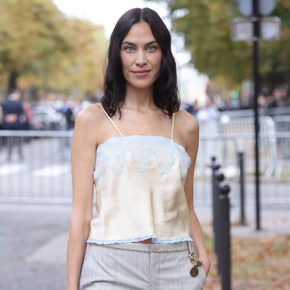 Alexa Chung poses for a photo outside of the Chloe SS27 runway show in Paris. She wears grey pinstripe trousers with a champagne lace-trimmed cami top and holds a beige Chloe Paddington bag.
