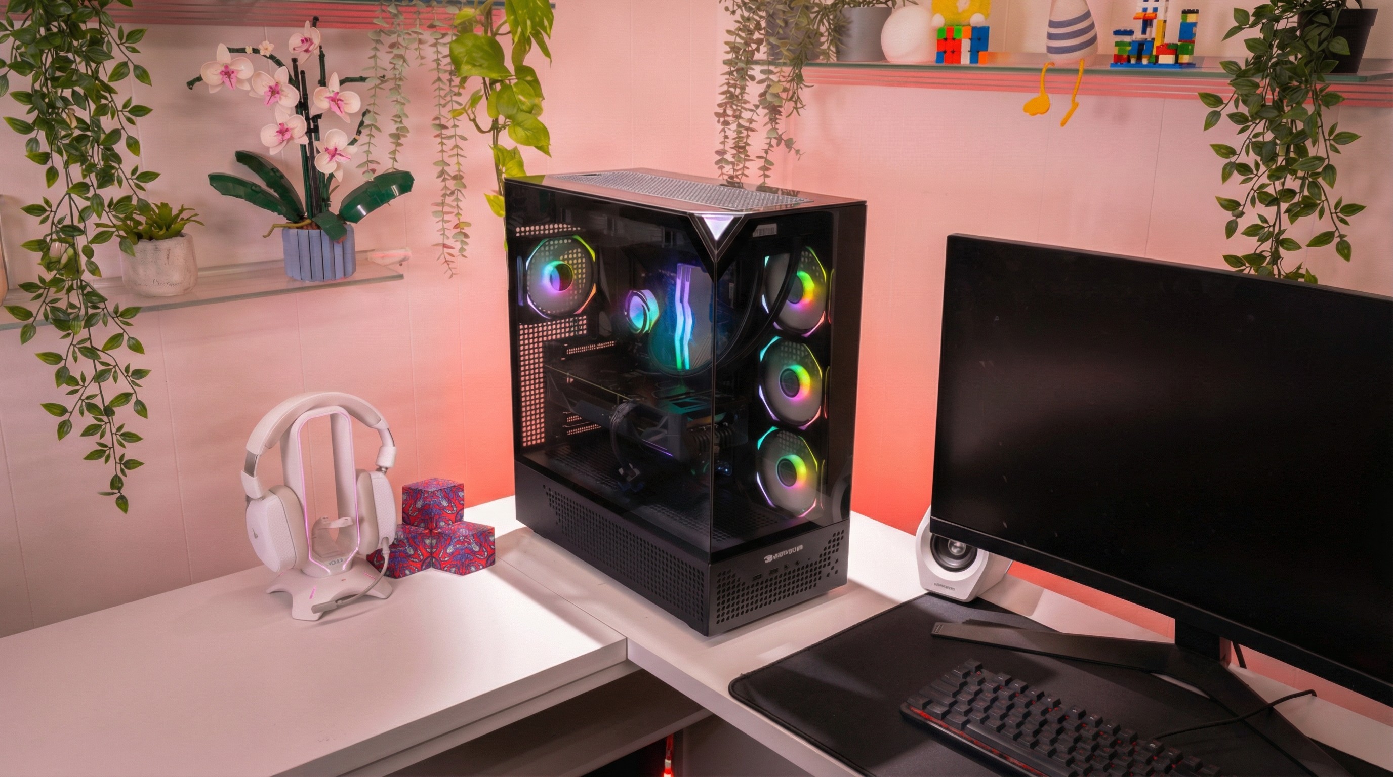 The iBuyPower RDY Element 9 Pro R07 Gaming PC on a white desk, next to a monitor, keyboard and headset, with leafy green plants trailing down from shelves above the desk and peachy pink lighting