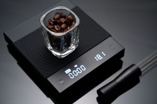 An espresso glass filled with coffee beans positioned on top of black coffee scales and a dark worktop