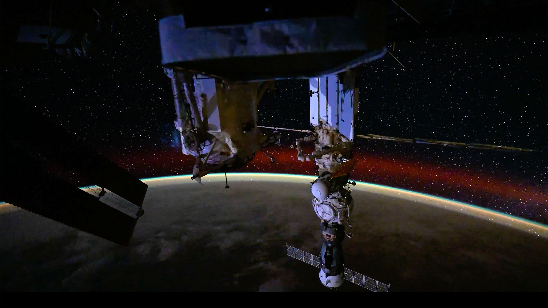 a docked spacecraft and space station modules are set against a starfield and the Aurora Australis (southern lights) above the horizon of Earth
