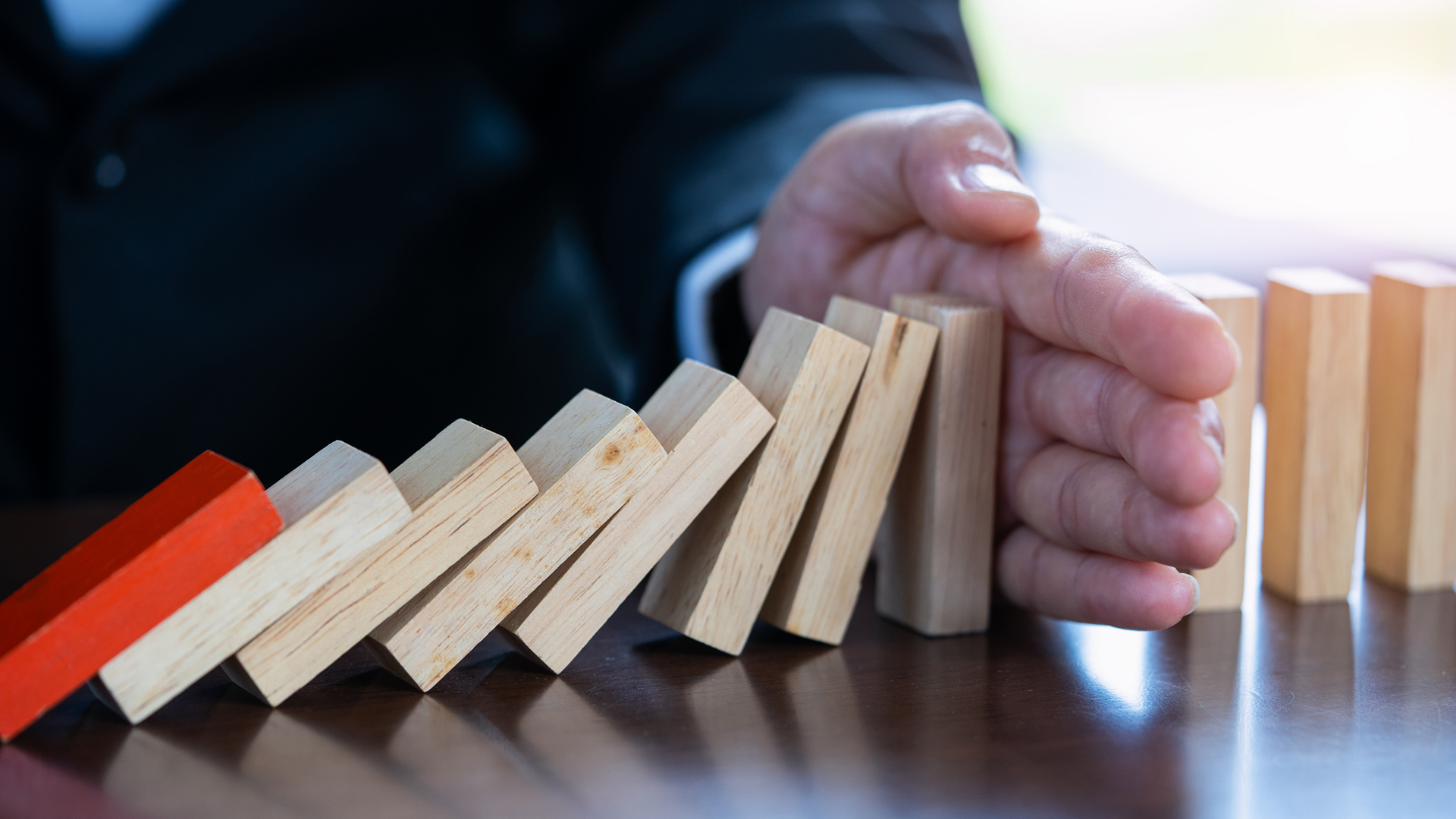A businessman's hand pushed in between wooden dominoes, stopping falling dominoes from knocking over others. The first domino that fell is red, while the rest are plain wood.