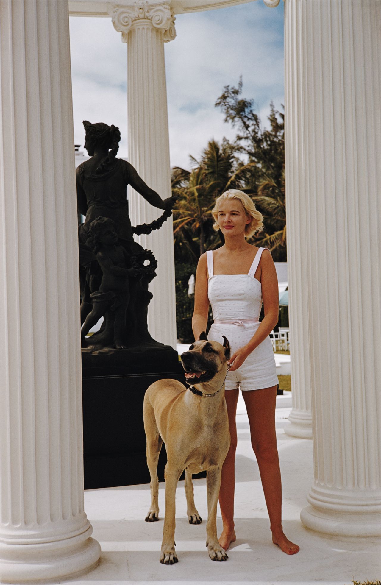 A woman in a 50s-style, white romper suit, holding on to the collar of a great Dane dog