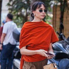 Woman wears velvet orange top, brown trousers, and flat shoes. 