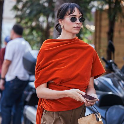 Woman wears velvet orange top, brown trousers, and flat shoes. 