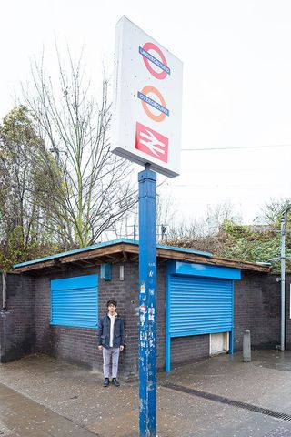 Outside image of Matthew Leung , stood at the Seven Sisters station small brick building shop, blue metal shutters closed, tall underground station sign with blue weathered post, concrete floor, trees and shrubs in the backdrop, grey sky