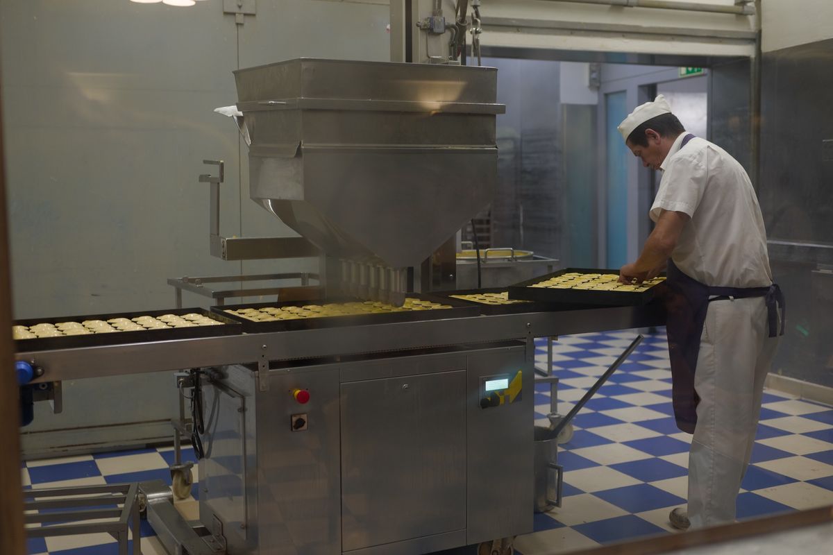 Baker in white uniform arranging pastries on trays beside industrial bakery machinery.