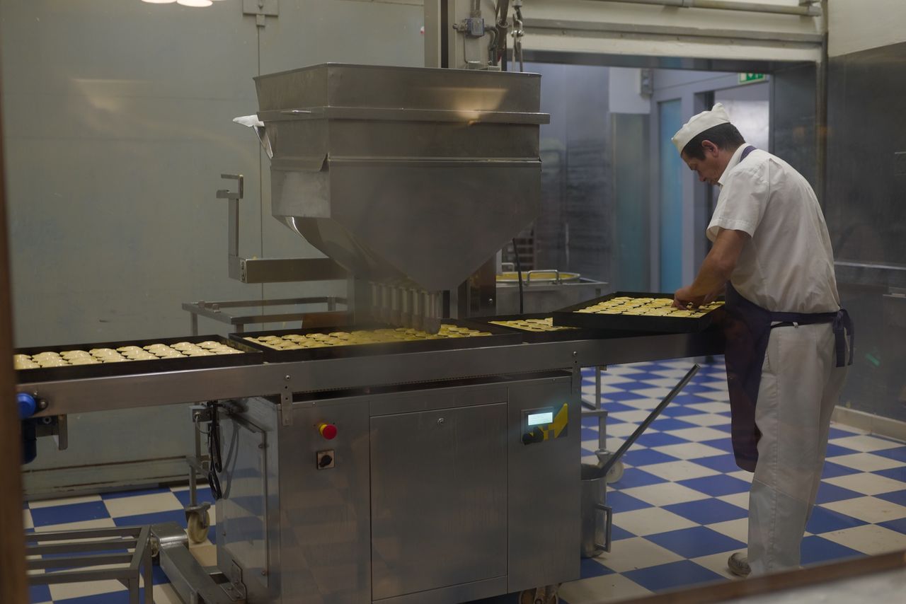 Baker in white uniform arranging pastries on trays beside industrial bakery machinery.