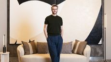 A young man with blonde hair, square brown glasses, a black T-shirt, and blue trousers stands in front of a large, monochromatic abstract painting and a beige couch with cushions surrounded by minimalist furniture.