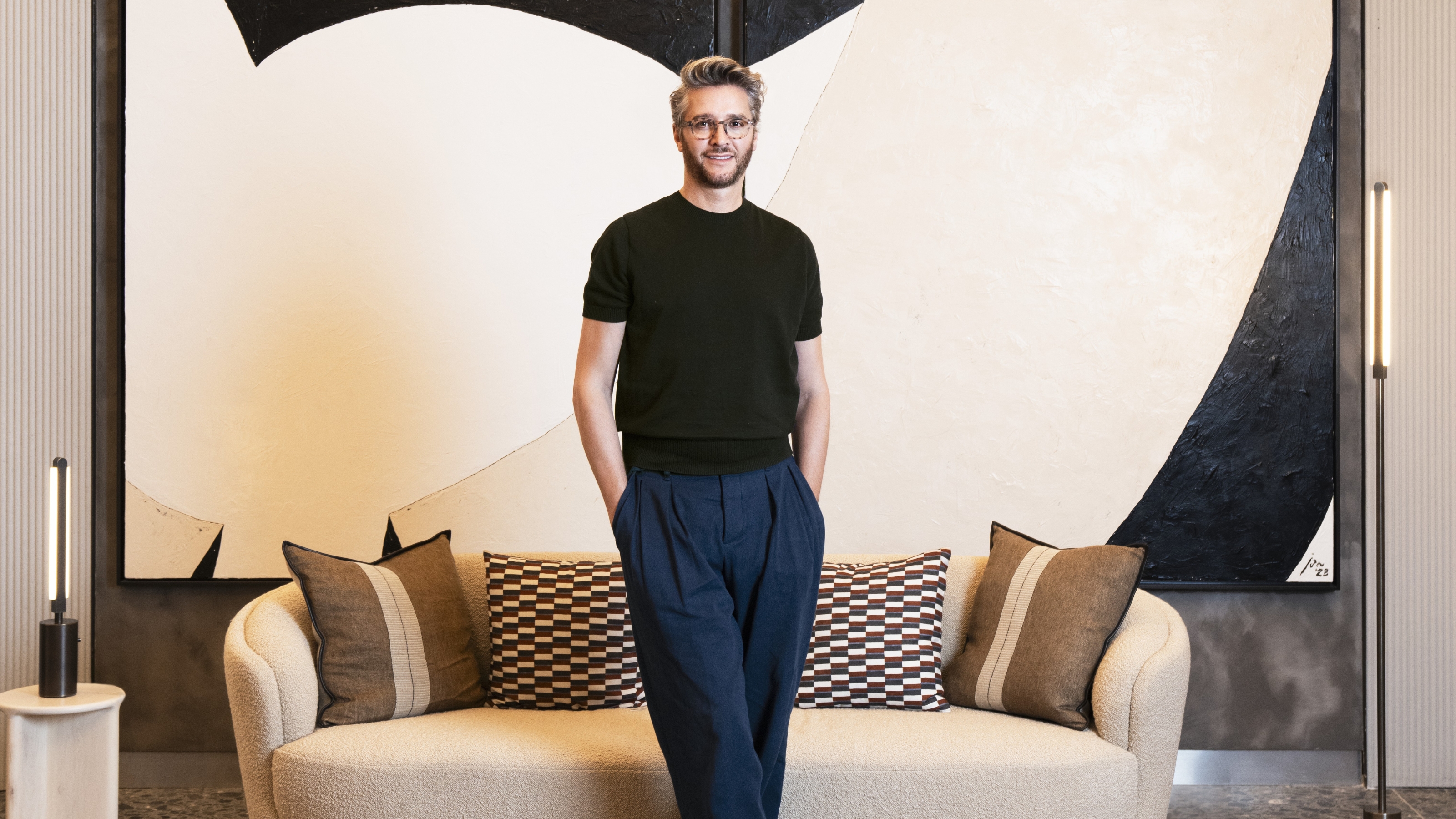 A young man with blonde hair, square brown glasses, a black T-shirt, and blue trousers stands in front of a large, monochromatic abstract painting and a beige couch with cushions surrounded by minimalist furniture.