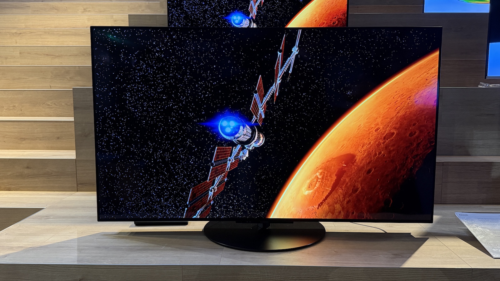 The Panasonic Z86C OLED TV pictured at a press event. On the screen is an image of a satellite orbiting an orange planet.