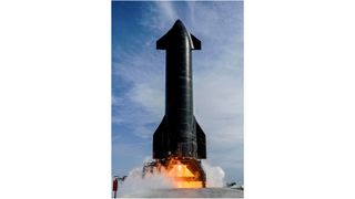 a large black rocket fires its engines on an outdoor test stand, with blue sky in the background