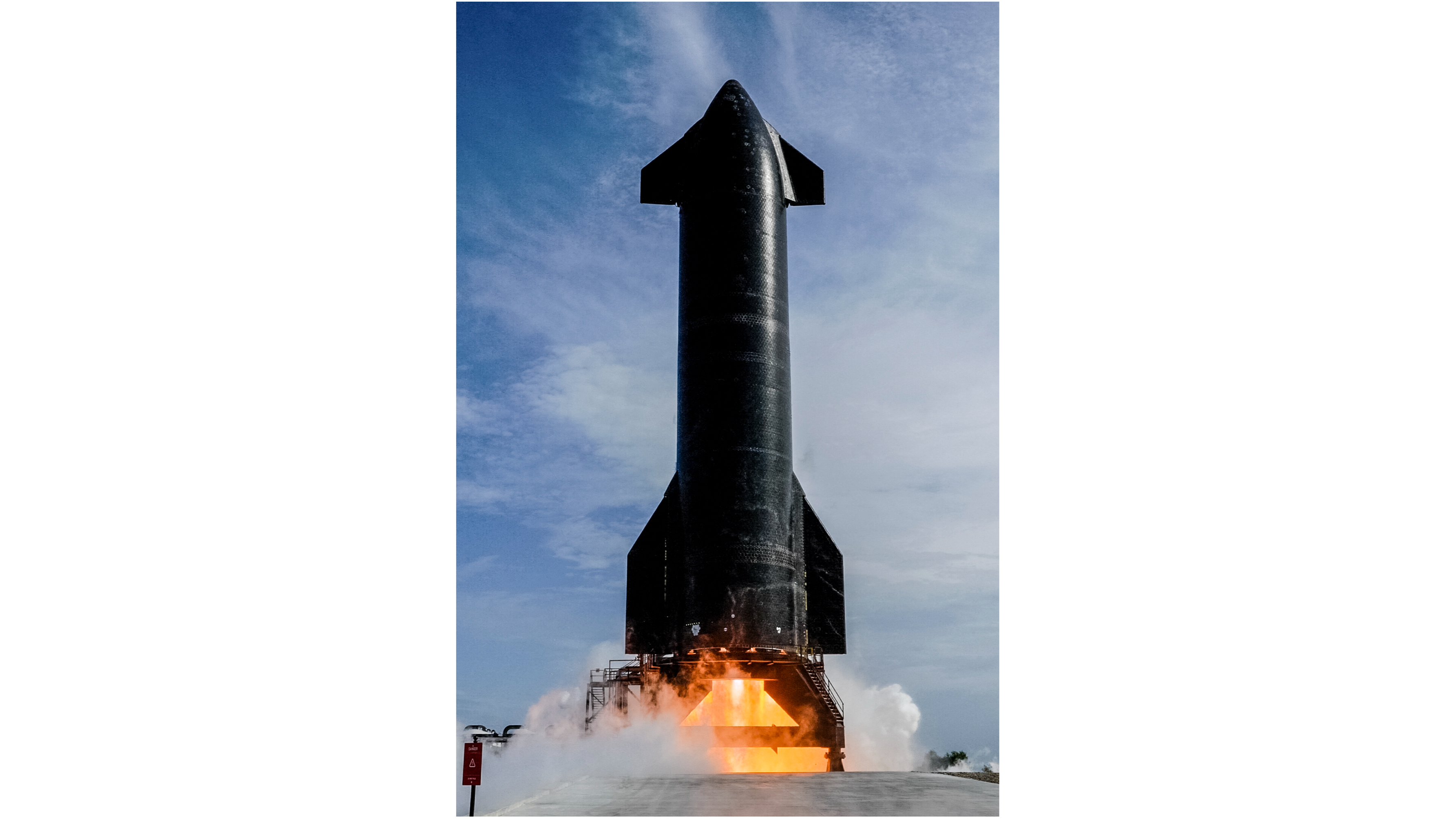 A large black rocket fires its engines on an outdoor test stand, with a blue sky in the background.