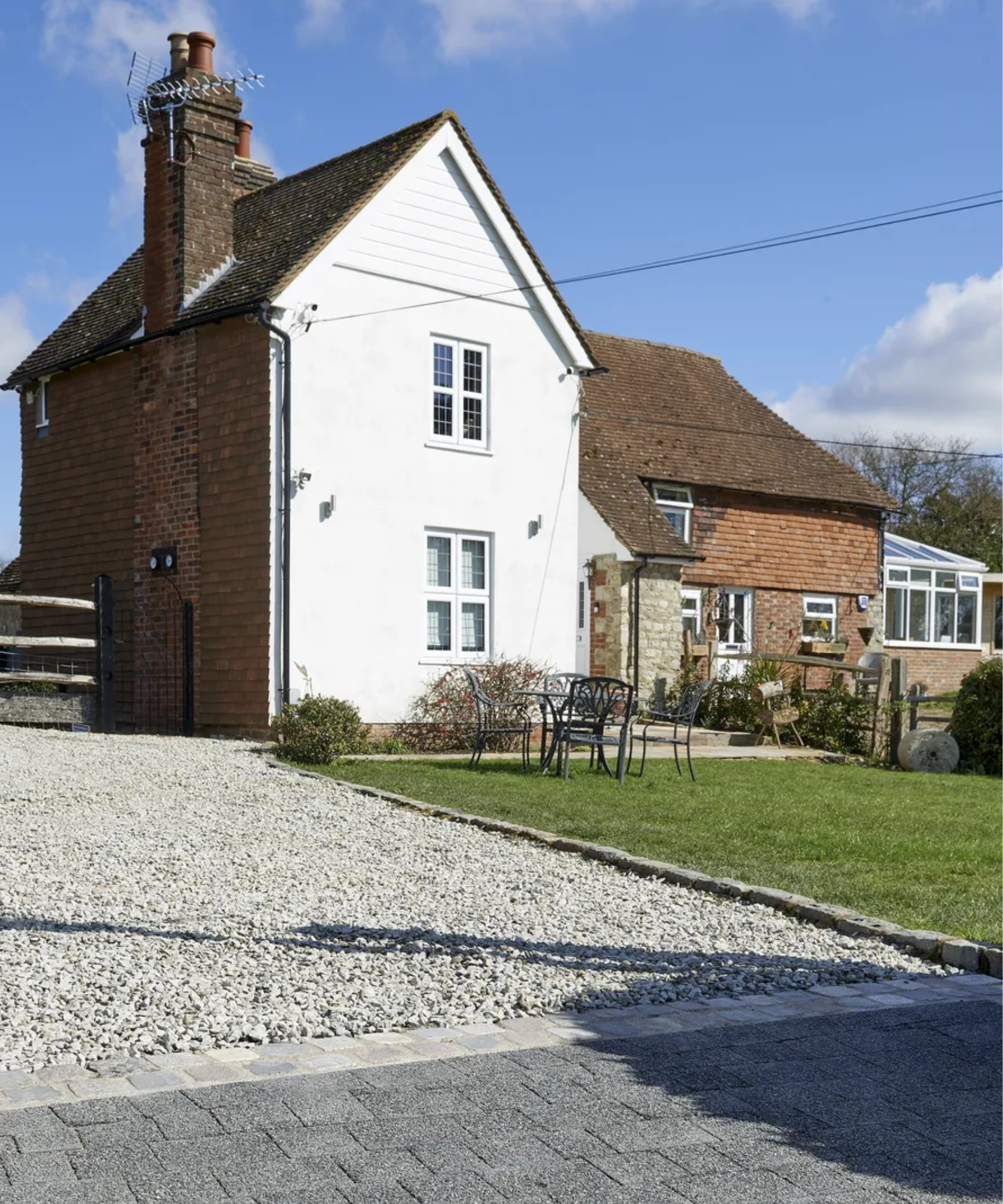 House with a gravel driveway to left, stones in the middle and a lawn on the right with outdoor chairs