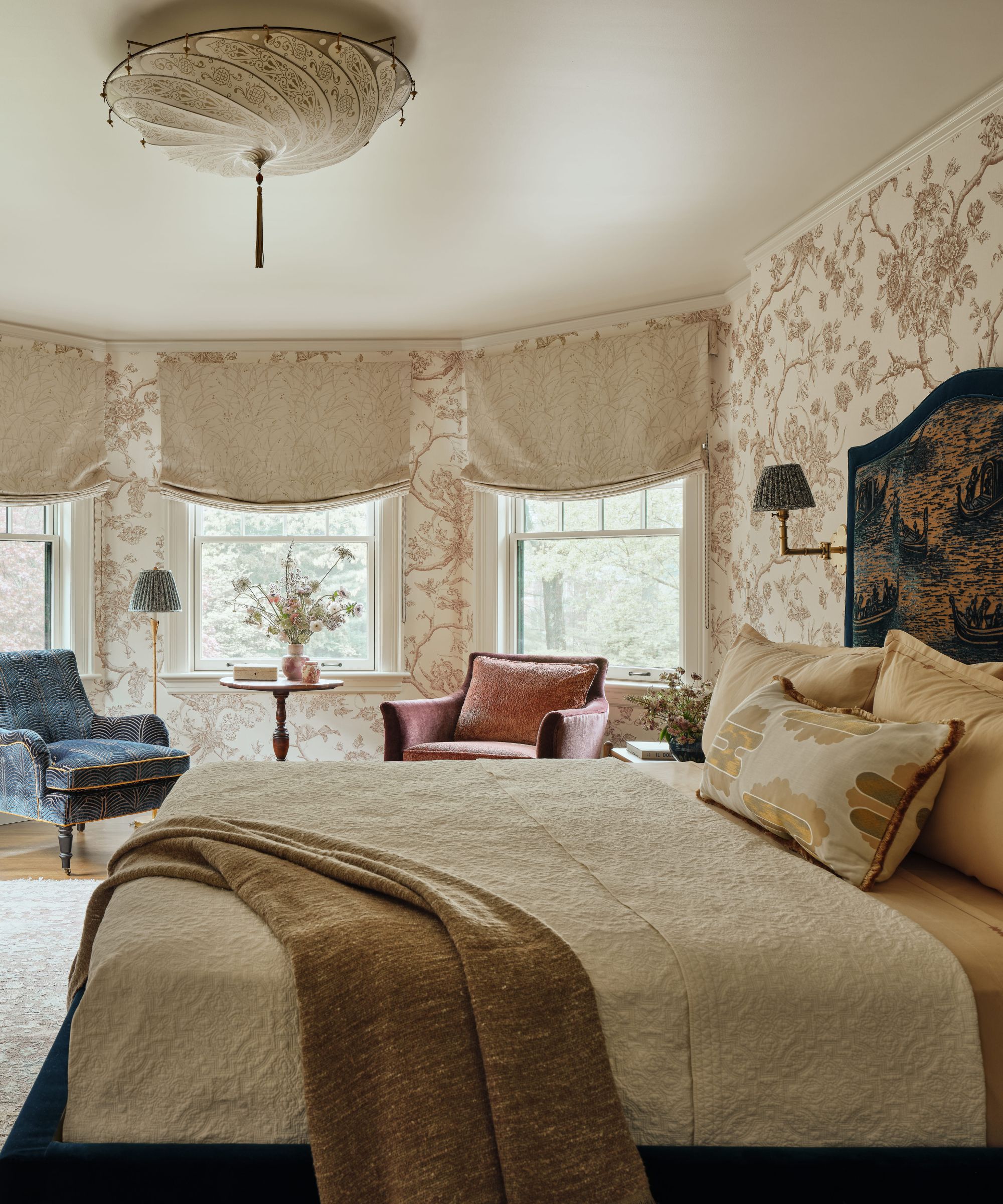 Bright bedroom with a cream-and-brown dressed bed, neutral floral wallpaper, and two patterned armchairs in a bay window.