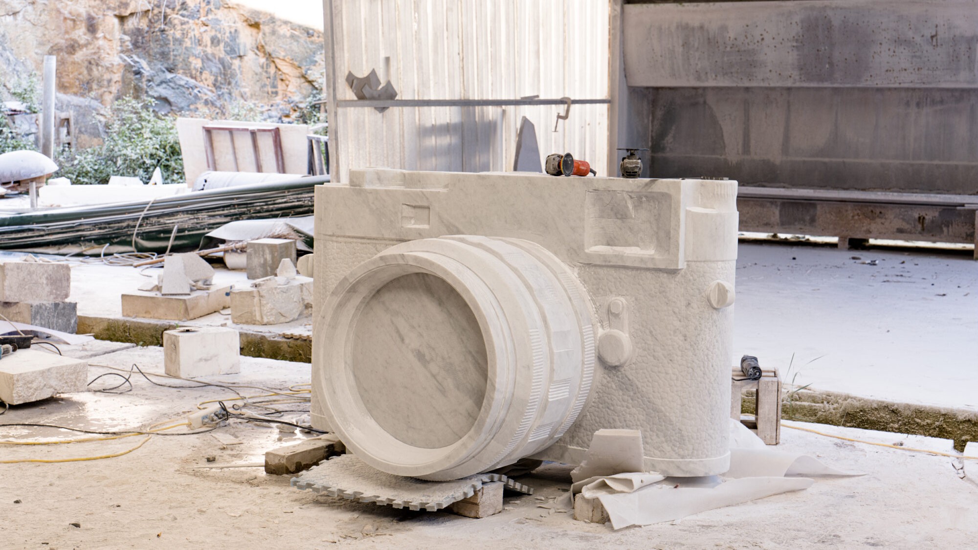 A large, monumental sculpture of a classic rangefinder camera, carved from white marble, sits on the dusty ground of an outdoor stoneworks or quarry workshop, surrounded by stone blocks and tools.
