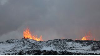 On a snowy mountain ahead, in the distance, two people stand ahead while lave spits out from the volcano in front of them.