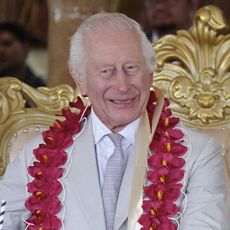 King Charles poses in a light gray suit and flowers smiling while Queen Camilla cries while wearing a blue and white floral tunic