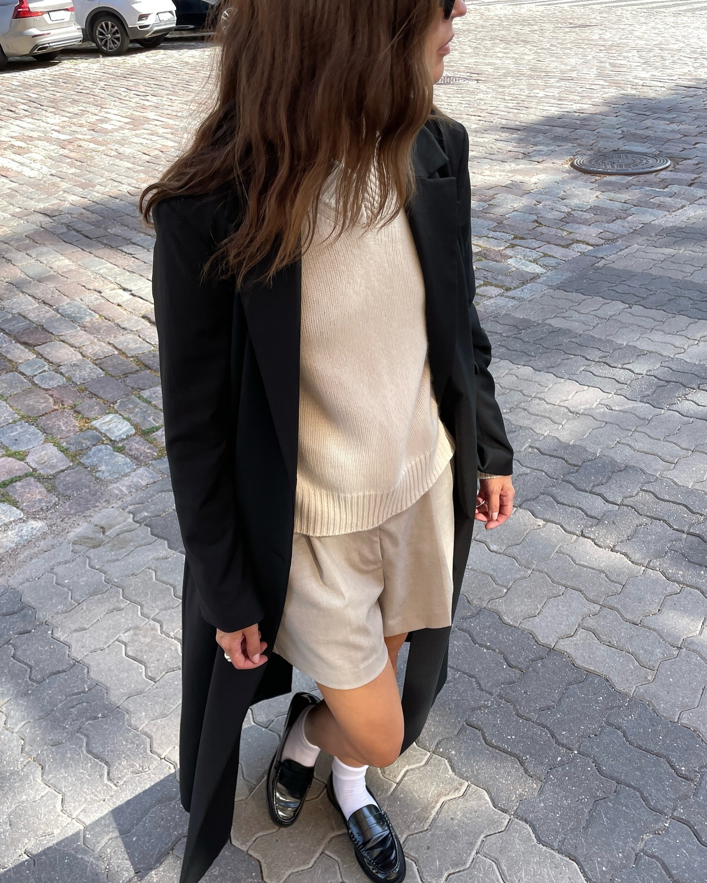 fashion influencer Johanna Piispa poses in a transitional spring outfit idea wearing a black maxi coat, beige V-neck sweater, beige shorts, white ankle socks, and black loafers