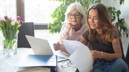 An older woman and her adult daughter look at paperwork together at the kitchen table.