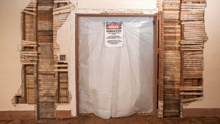 part exposed lath and plaster walls with opening into another room taped over with plastic and asbestos warning sign