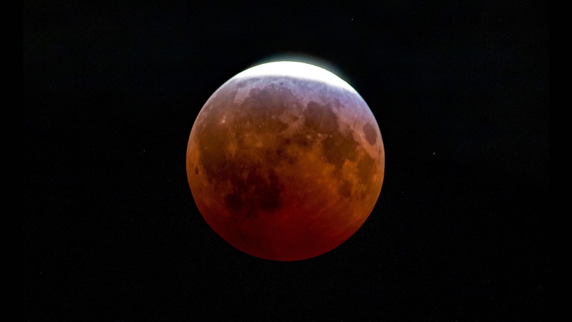 An red, eclipsed full moon is photographed in a black sky with a thin crescent at the top exposed to direct sunlight.