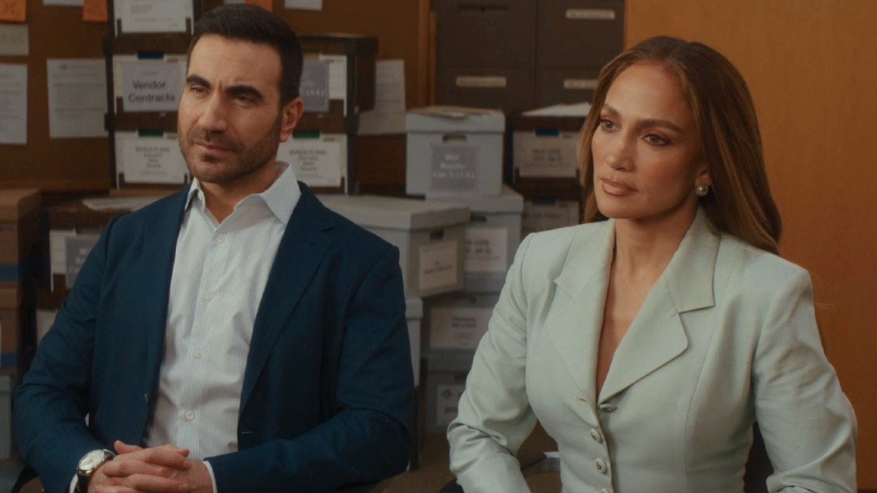 From left to right: Brett Goldstein and Jennifer Lopez sitting next to each other in an office in Office Romance. 