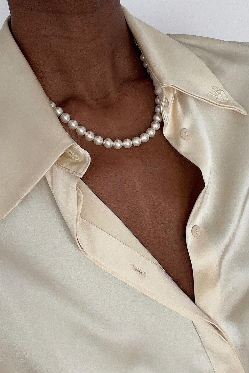 a woman wearing a cream satin button down, and white pearl necklace