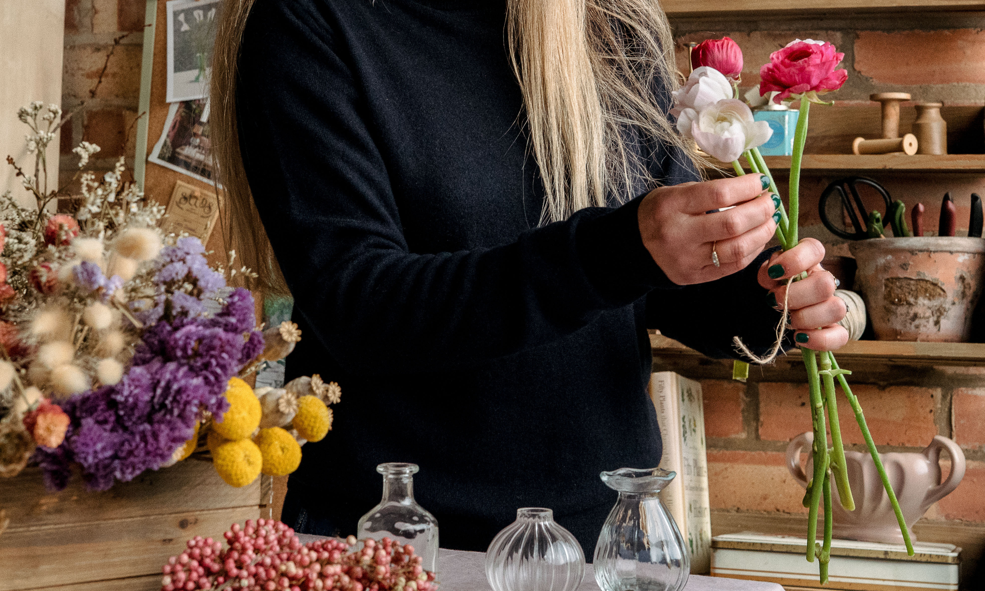 Hands holding fresh, pink ranunculus flowers, with a selection of dried flowers on the bench below