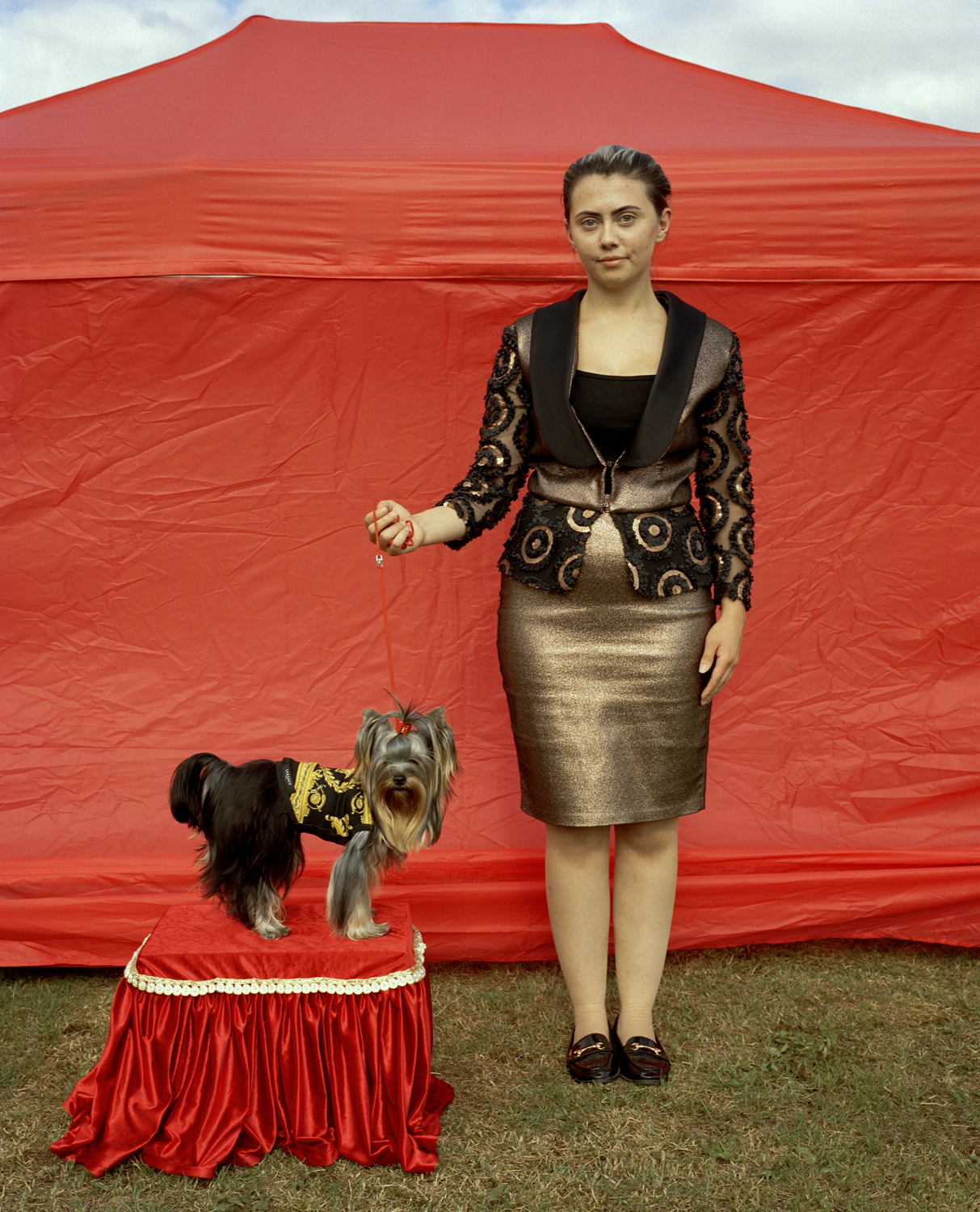 A young woman in a gold lam&amp;eacute; skirt and ornate beaded jacket holds the lead of a Yorkshire Terrier posed on a red satin plinth, both standing before a vivid red pop-up tent.