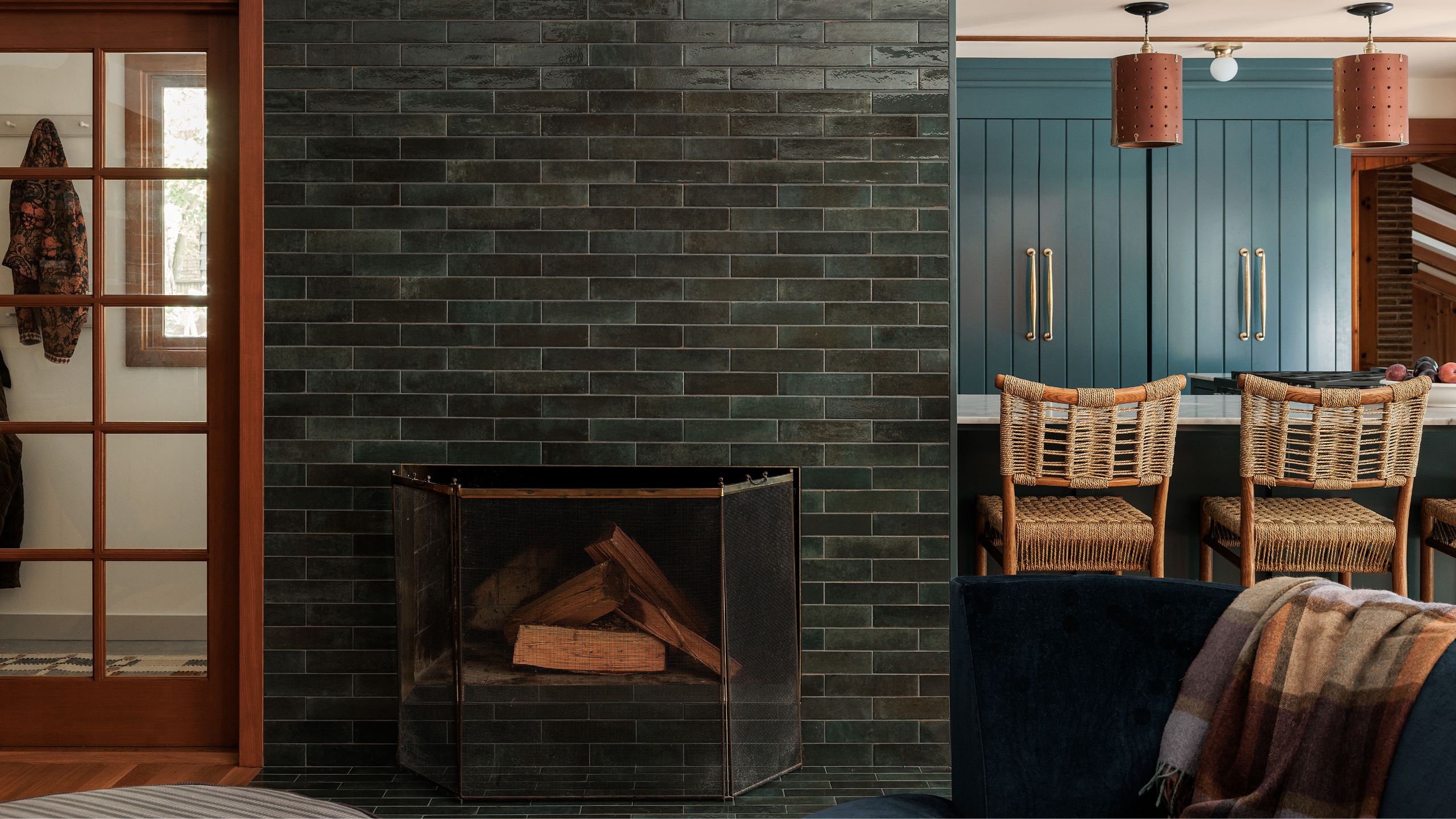 a cozy entry with green tiled fireplace, a wood glass door and view through to a blue green kitchen with island and rattan bar stools