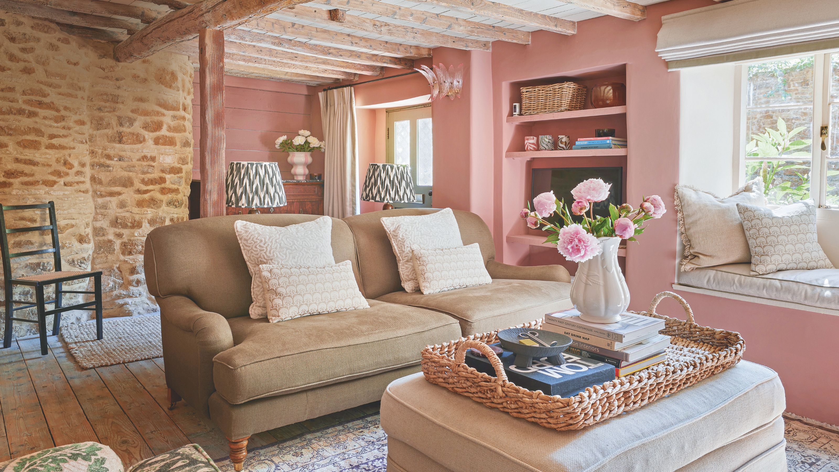Pink cottage living room with exposed brick and beams, cosy sofa, wicker tray on footstool, and window seat