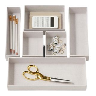 A set of six beige material drawer organizers filled with white and gold pencils, scissors, paperclips and a white calculator on top of a brown notebook