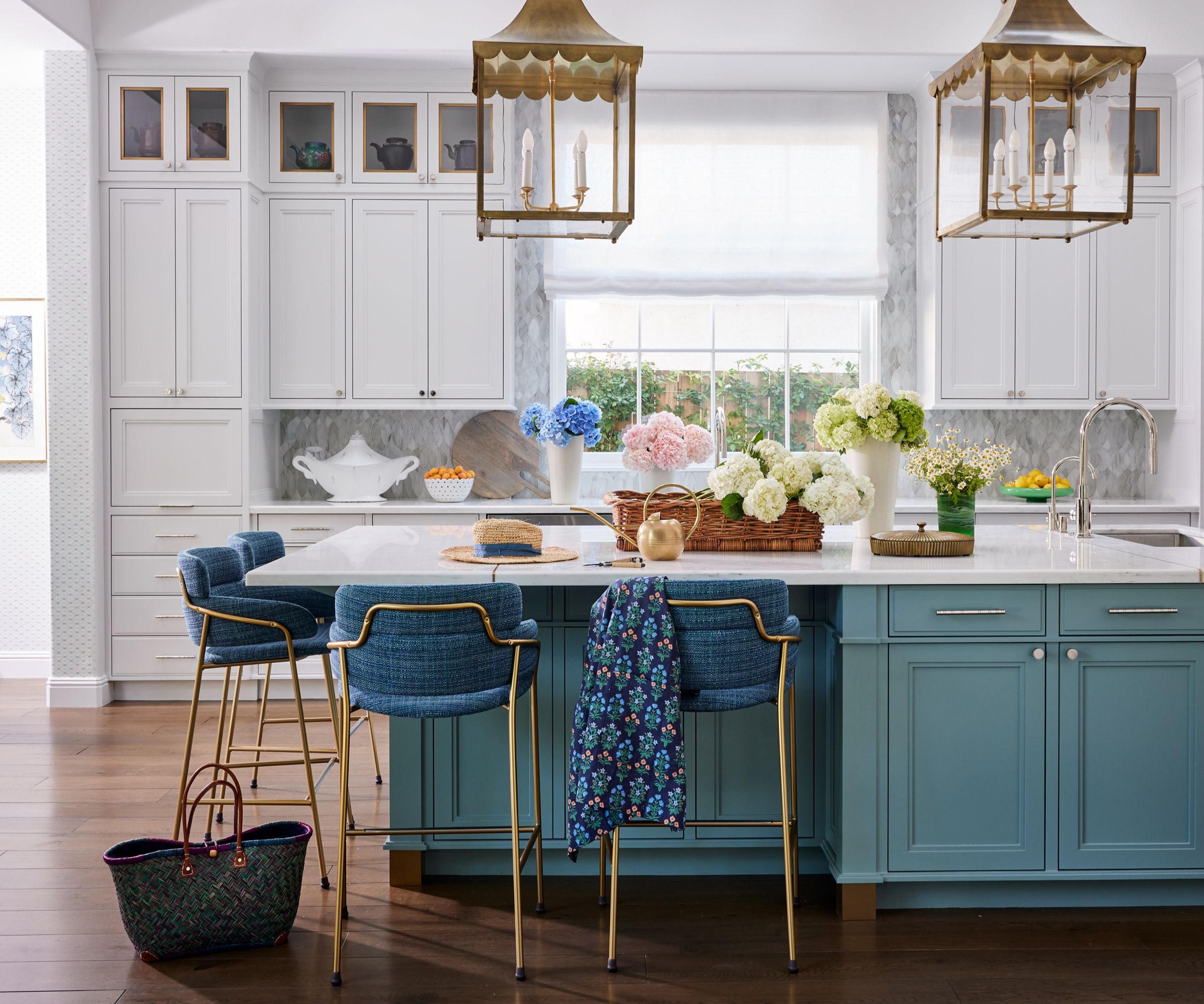 A spacious kitchen with a central island painted in teal blue, featuring gold and blue bar stools. The cabinetry is white with gold hardware, and the backsplash is a soft gray. Two large gold pendant lights hang over the island