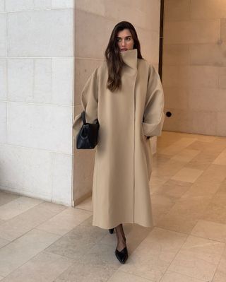 Woman wearing nude funnel-neck coat, black heels, and black bag.