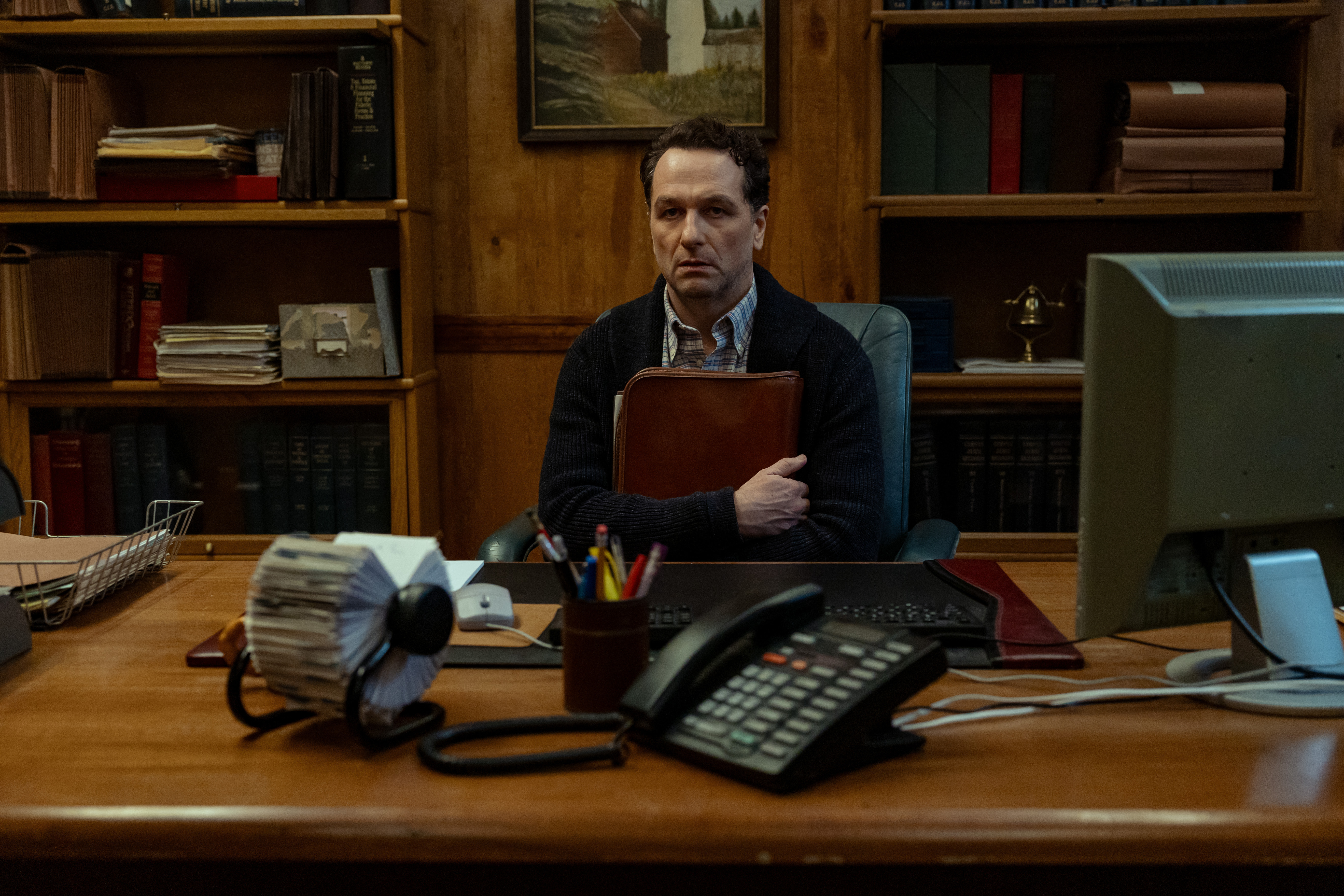 Matthew Rhys as Mayor Tom Loftis, looking scared while holding a red book to his chest and sitting behind an office desk, in the Apple TV show #039;Widow#039;s Bay.#039;