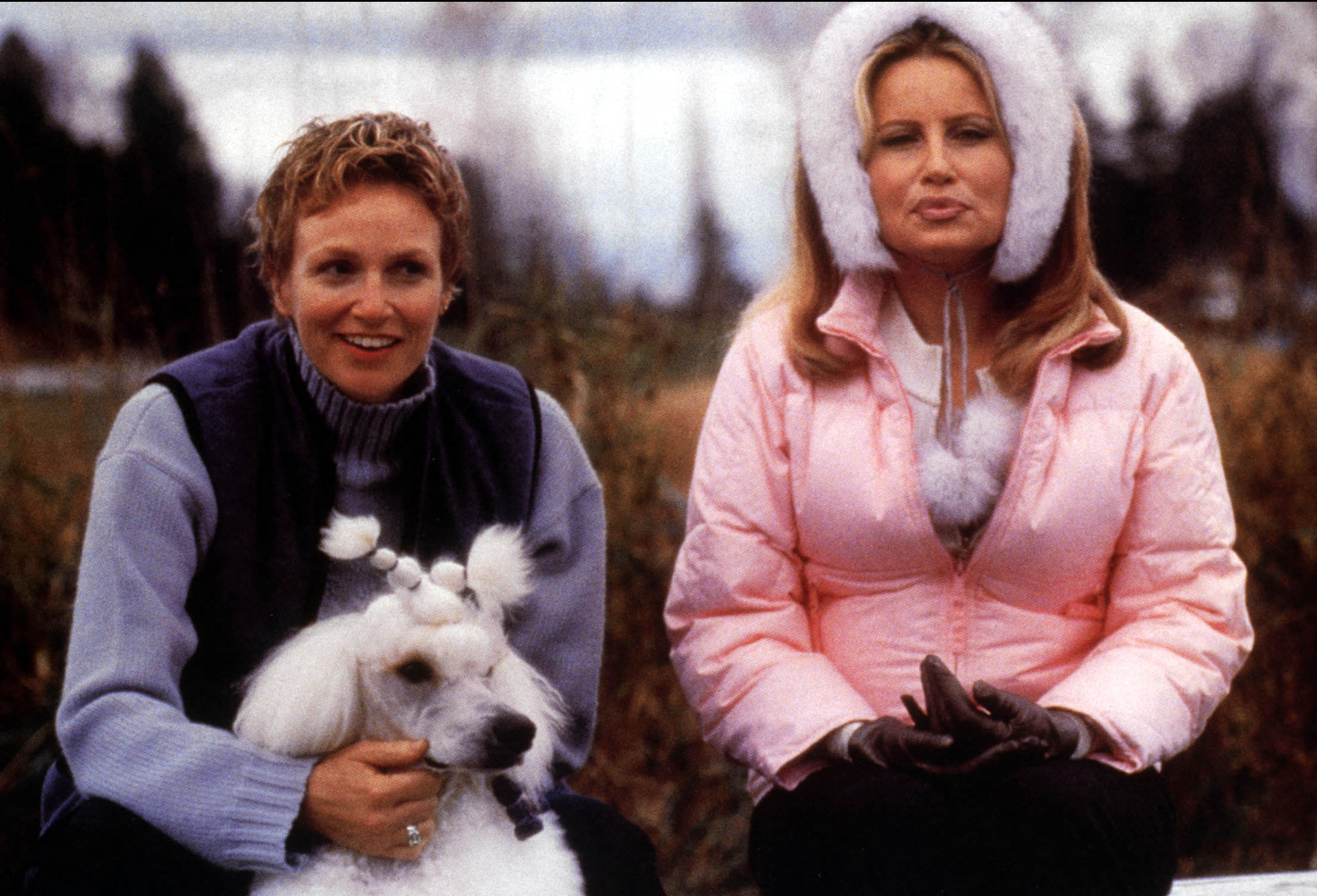 Two women sit outdoors in cold weather—one holding a white poodle groomed with pom-poms, the other wearing a pink puffer coat with fur-trimmed ear muffs.