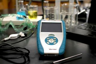 The Talking LabQuest, on a laboratory bench next to goggles, ear buds, beakers and a computer. The temperature probe is plugged in and is visible in the foreground.