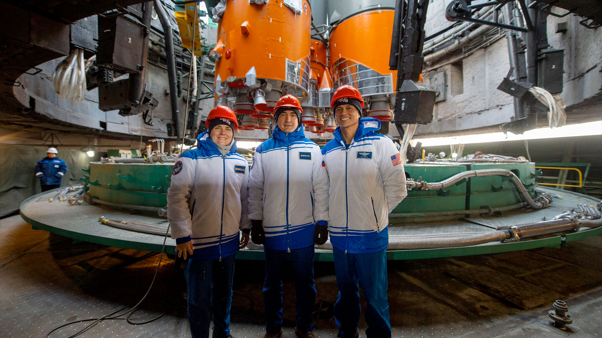 two men and a woman (at left) wearing white and blue jackets and red hard hats pose for a photo at the base of a rocket on a launch pad.