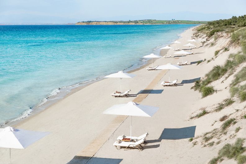 A section of the unspoilt Bousoulas Beach at Sani Resort. There are soft sand dunes with tuffs of grass, soft white sand, turquoise see and white parasols and loungers dotted along the beach