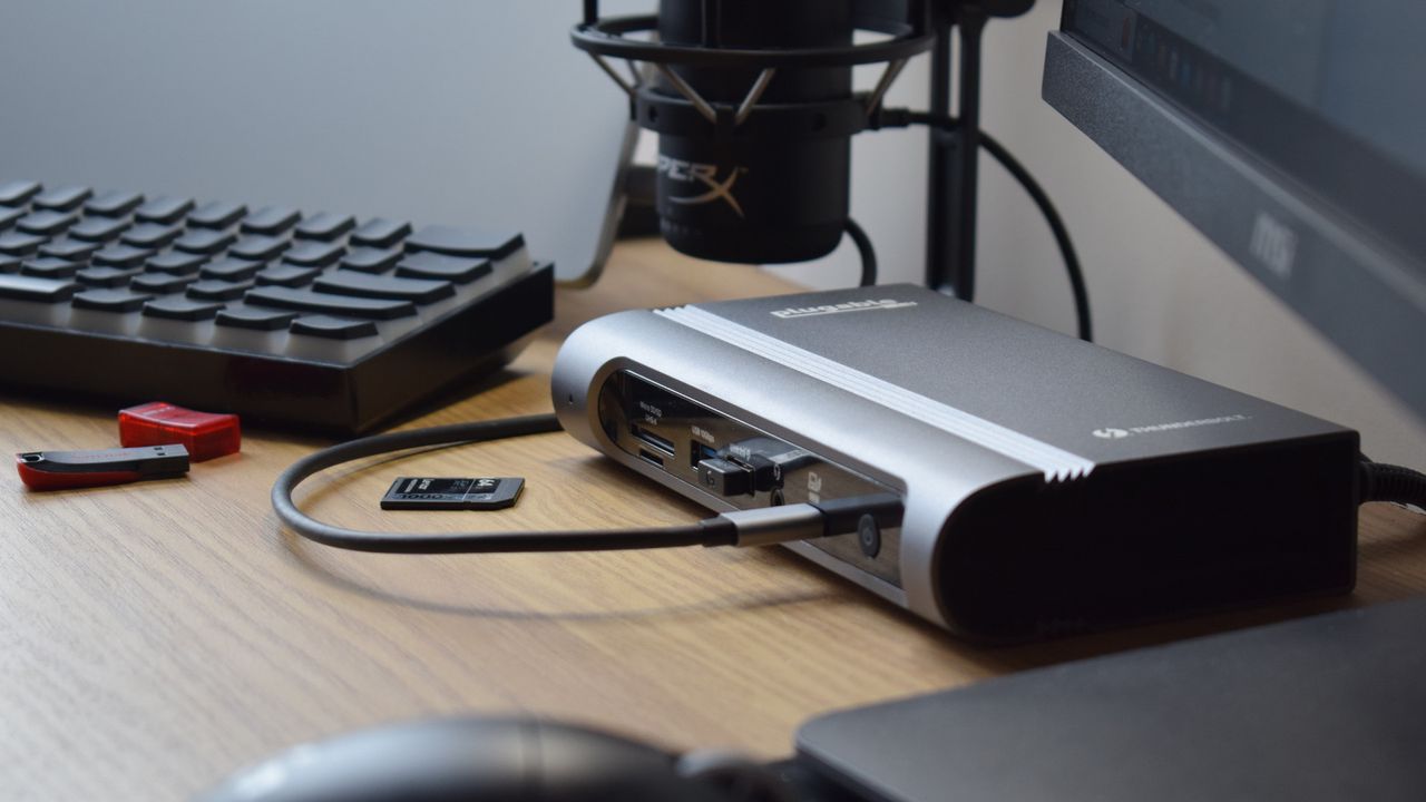 Photo of the Plugable Thunderbolt 4 &amp; USB4 Quad Display Docking Station (TBT4-UDZ) as part of a desk setup connected to a laptop, microphone, external display, and wireless keyboard.