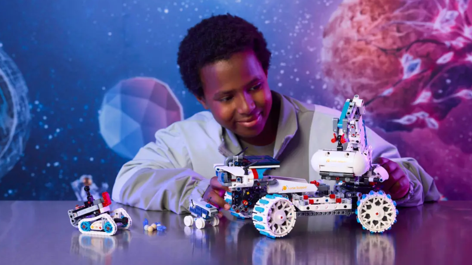 A child in a space-themed room smiles at a white Lego rover, which is sat on a reflective surface