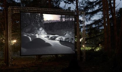 Dusk image, green and brown forest area, metal frame suspending a projector screen showing a black and white image of a snowy forest, stream and waters edge of crisp white snow, ground spotlights shining behind the metal frame, clear sky